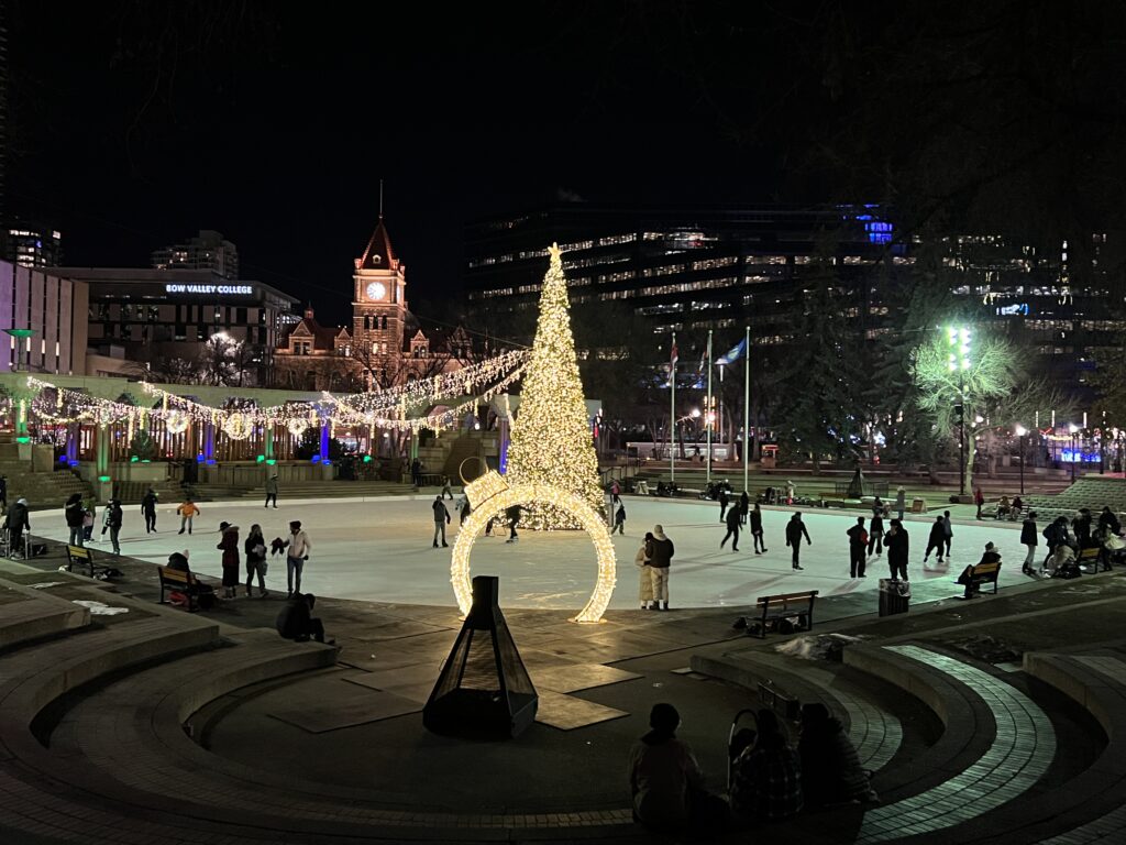 Image of a tall lit up Christmas tree in the center of an iceskating rink in Olympic Plaza- Calgary 